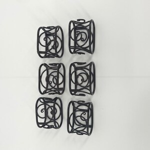 May include: A set of six black metal napkin rings arranged in two columns of three. Each ring features a cylindrical shape constructed from thin, curved wire forming an openwork scroll pattern with circular and swirling decorative elements.