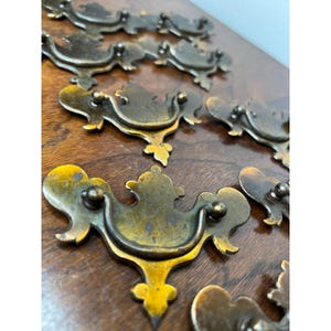 Puede incluir: Una colección de tiradores de cajón de metal en tono latón con un asa curva oscilante unida a una placa decorativa con bordes ornamentados y simétricos. Los herrajes presentan un acabado envejecido con pátina y manchas oscuras.