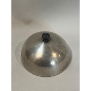 May include: A silver-colored, dome-shaped cloche with a small, dark knob on top. The cloche has a smooth, reflective surface and is likely made of metal. It is designed to cover and protect food items.
