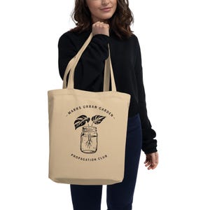 May include: Beige tote bag with black text and a plant propagation design. The bag features the text "MARKS URBAN GARDEN" above a jar illustration with leaves and roots, and "PROPAGATION CLUB" below. The bag is held by a person wearing a black sweater.