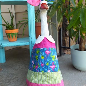 May include: A decorative goose dressed in a colorful outfit. The goose wears a pink and blue patterned dress with a white collar and matching headpiece. The dress features a flamingo and palm tree design.