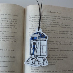May include: A white and blue embroidered bookmark featuring a Star Wars character, R2-D2. The bookmark is attached to a black string. The text "Retusa means death. Kill me then. I am your Prince and I'm not that bad-how could you rather be" is embroidered on the bottom of the bookmark.