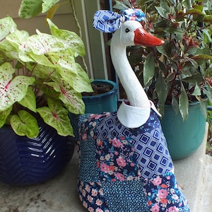May include: A decorative goose statue with a white head and neck, a red beak, and a blue bow. The goose wears a patchwork dress with floral and geometric patterns. Potted plants in blue pots are in the background.