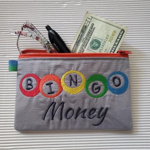 May include: A gray zippered pouch with an orange zipper featuring embroidered colorful circles containing the letters B, I, N, G, O and the word Money in dark thread. Inside are a black pen, eyeglasses, and a twenty-dollar bill.