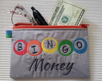 May include: A gray zippered pouch with an orange zipper featuring embroidered colorful circles containing the letters B, I, N, G, O and the word Money in dark thread. Inside are a black pen, eyeglasses, and a twenty-dollar bill.
