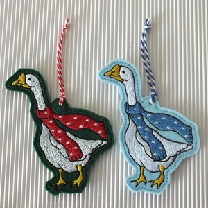 May include: Two felt goose ornaments with embroidered details. One goose is wearing a red and white polka dot scarf and is on a green background. The other goose is wearing a blue and white polka dot scarf and is on a light blue background.
