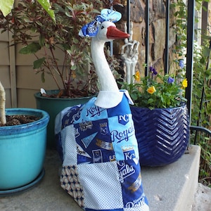May include: A decorative goose statue with a white neck and head, a red beak, and a blue and white patterned outfit. The outfit features the word "Royals" and baseball-themed designs. A matching bow adorns the goose's head. The goose is displayed outdoors near potted plants.