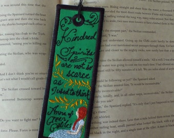 Könnte beinhalten: Grünes Lesezeichen mit gesticktem Text, der "Kindred Spirits are not so scarce as I used to think" und "Anne of Green Gables" lautet. Das Lesezeichen hat einen grünen Hintergrund mit goldener und weißer Stickerei. Das Lesezeichen ist in der Mitte gefaltet und liegt auf einem Buch.