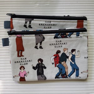 May include: Two rectangular, zippered pouches with a light gray background. The pouches feature a repeating pattern of figures from "The Breakfast Club" movie and the text "THE BREAKFAST CLUB". The zippers are black.
