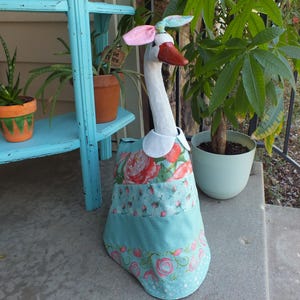 May include: A whimsical goose statue with a white neck and head, a red beak, and pink ears. It wears a blue dress with a floral pattern and a white collar. The statue is outdoors near potted plants.