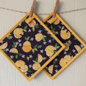 May include: Two quilted lemon-themed pot holders with a yellow border and white polka dots. The pot holders have a dark purple background with a pattern of lemons, leaves, and white flowers.