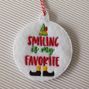 May include: White felt Christmas ornament with red and green embroidery. The ornament features an elf hat and the text "Smiling is my favorite".