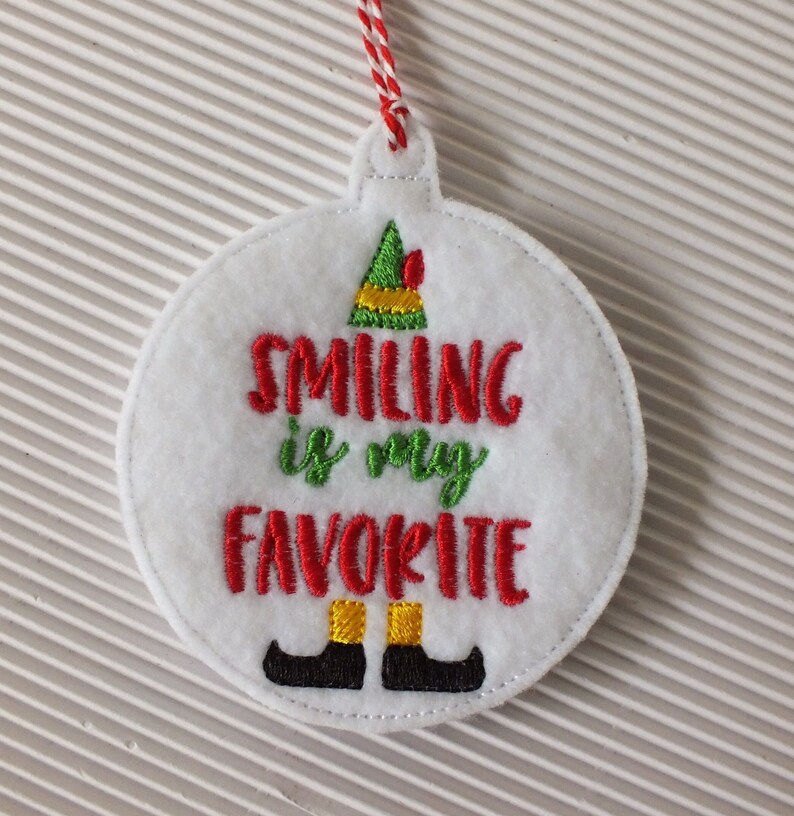 May include: White felt Christmas ornament with red and green embroidery. The ornament features an elf hat and the text "Smiling is my favorite".