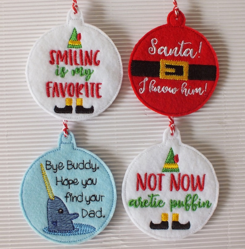 May include: Four felt Christmas ornaments with embroidered designs. The first ornament is white with green and black embroidery that says "Smiling is my favorite". The second ornament is red with black and gold embroidery that says "Santa! I know him!". The third ornament is light blue with blue and yellow embroidery that says "Bye Buddy. Hope you find your Dad." The fourth ornament is white with green, black, and yellow embroidery that says "Not Now arctic puffin".