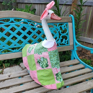 May include: A decorative goose figurine wearing a pink and green patchwork fabric cover with a succulent print and white polka dots. The goose has a white collar around its neck and a pink fabric bow tied on top of its head, featuring an orange beak.