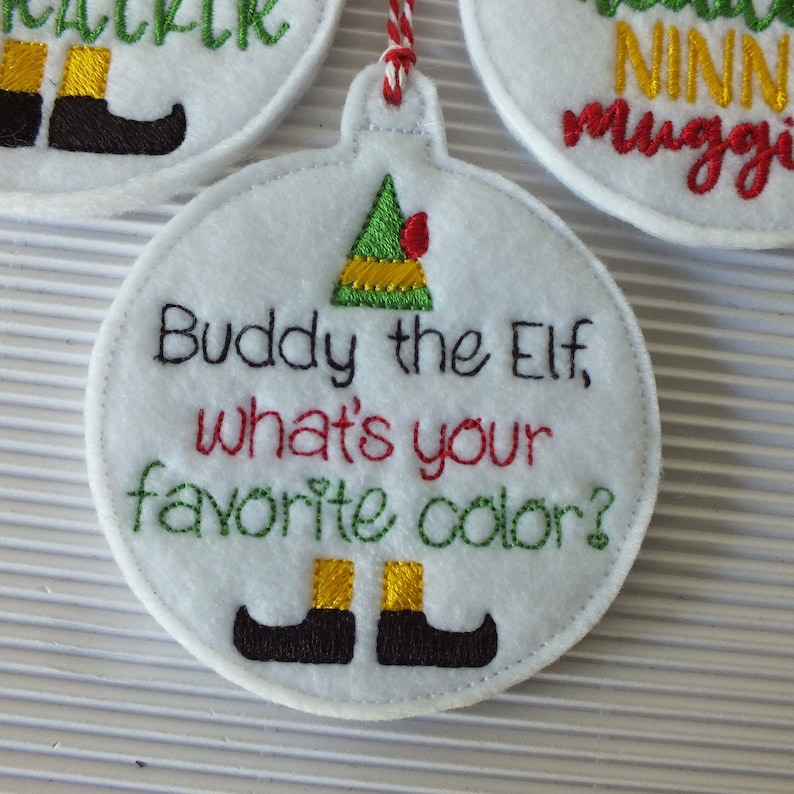 May include: White felt Christmas ornament with green, yellow, red, and black embroidery. The ornament features an elf with the text "Buddy the Elf, what's your favorite color?"