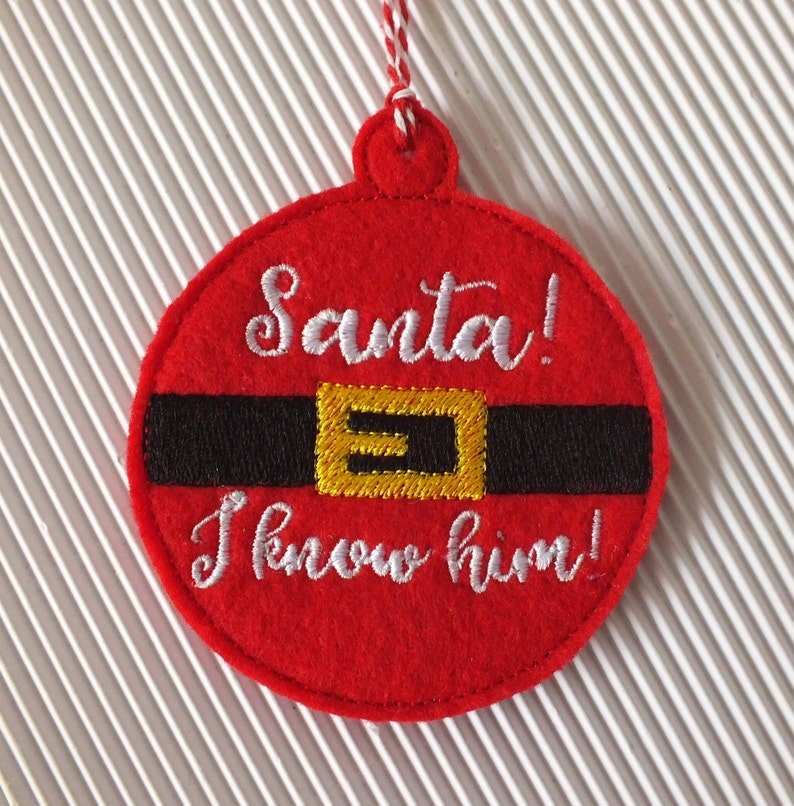 May include: Red felt Christmas ornament with white embroidery that reads "Santa! I know him!" and a black and gold belt design.