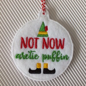 May include: White felt Christmas ornament with green, red, and yellow embroidery. The ornament features an elf hat and the text "NOT NOW arctic puffin".