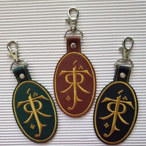 May include: Three keychains with oval-shaped pendants. The pendants are dark green, brown, and black, each featuring a gold embroidered design. Each keychain has a silver-colored clasp and a leather strap.