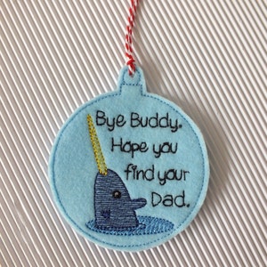 May include: A blue felt ornament shaped like a Christmas tree ornament with a narwhal embroidered on it. The narwhal has a gold horn and black eyes. The ornament has the text "Bye Buddy. Hope you find your Dad." embroidered on it.