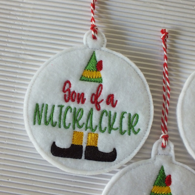 May include: White felt Christmas ornament with red and white twine. The ornament features an embroidered elf with the text "Son of a Nutcracker".