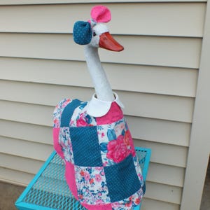 May include: A whimsical goose figurine with a white neck and head, a red beak, and a patchwork quilt-style dress. The goose wears a blue bow and a pink bow on its head. The dress features pink, blue, and floral patterns.