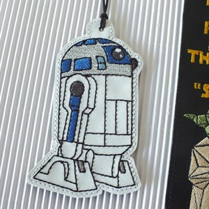 May include: White and blue embroidered R2-D2 bookmark with a black string attached. The bookmark is shaped like the Star Wars character R2-D2.