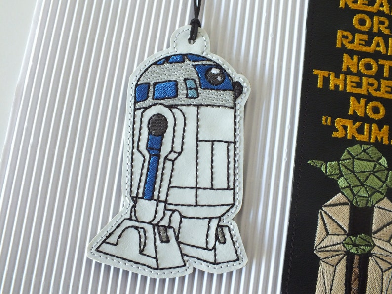 May include: White and blue embroidered R2-D2 bookmark with a black string attached. The bookmark is shaped like the Star Wars character R2-D2.