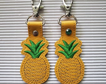 May include: Two yellow pineapple keychains with green embroidered leaves and silver-tone clasps. The keychains are made of leather and have a stitched design. One keychain has a green gem.