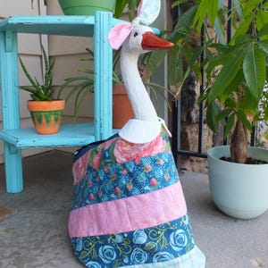 May include: A whimsical goose statue with a white neck and head, a red beak, and pink and blue ears. It wears a colorful patchwork dress with floral and striped patterns. The statue is a decorative garden accent.