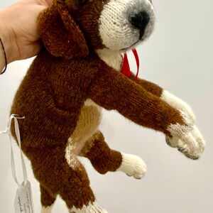 Könnte beinhalten: Handgestricktes Hundespielzeug in Braun und Weiß. Der Hund hat Schlappohren, einen weißen Streifen auf dem Kopf und ein rotes Band. Die Beine und Pfoten sind weiß. Ein Etikett ist am Spielzeug befestigt.