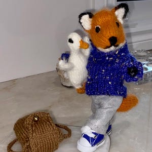 May include: A handmade knitted fox doll with orange fur, wearing a blue sweater, gray pants, and blue sneakers. The fox is holding a white duck doll. A brown knitted backpack is on the surface.