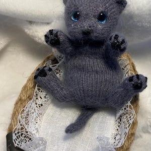 Könnte beinhalten: Ein graues, gestricktes Katzenspielzeug mit blauen Augen, das in einem geflochtenen Korb sitzt, der mit weißem Stoff und Spitze ausgekleidet ist.