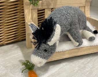 May include: A knitted gray donkey toy with a black mane, white muzzle, and white hooves, positioned on a small wooden bench. The donkey is facing downward toward a small, knitted orange carrot with green stems placed on the surface below.