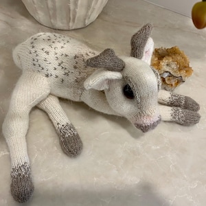 Op de afbeelding: Een handgemaakt gebreid hertenspeelgoed in crème en grijs. Het hert ligt met uitgestrekte poten. Het heeft bruine knoopogen, grijze geweien en grijze hoeven. Het lichaam van het hert heeft een patroon van kleine grijze pijlen.