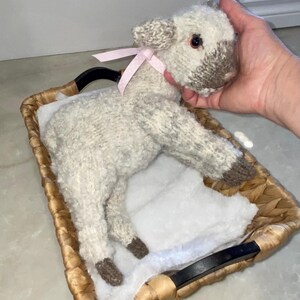 Könnte beinhalten: Ein handgestricktes Lammspielzeug mit rosa Schleife, das in einem geflochtenen Korb liegt. Das Lamm ist hauptsächlich weiß mit grauen Akzenten an Kopf und Füßen. Der Korb hat schwarze Griffe und ein weißes, flauschiges Innenfutter.