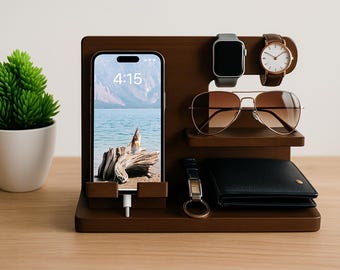May include: A wooden desk organizer with a smartphone displaying a landscape image, a smartwatch, a wristwatch, sunglasses, a wallet, and a keychain. A small potted plant is on the left. The time on the phone is 4:15.