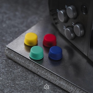 May include: Four small, cylindrical, matte-finish buttons in yellow, red, teal, and blue are arranged on a dark metallic surface. Next to them is a black device featuring four circular silver buttons labeled with the letters A, B, H, and L.