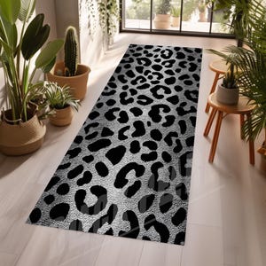 May include: A rectangular rug with a silver background and black leopard print pattern. The rug is placed on a light-colored wooden floor, surrounded by potted plants and natural light.