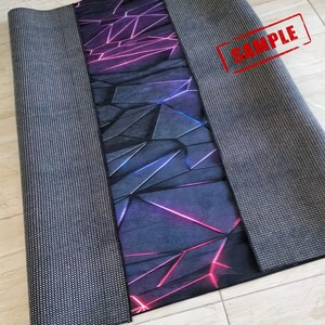 Puede incluir: Una alfombra rectangular con un dise&ntilde;o geom&eacute;trico en tonos morado, azul y rosa. El patr&oacute;n est&aacute; compuesto por formas angulares con l&iacute;neas brillantes. La alfombra tiene un borde gris oscuro y un reverso antideslizante.