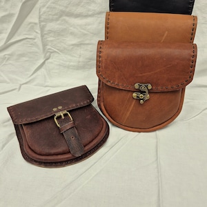May include: Three leather pouches with different colored leathers. The top pouch is black, the middle pouch is brown, and the bottom pouch is dark brown. The pouches have a flap closure with a metal buckle.