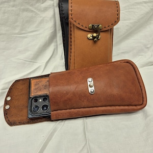 May include: Two leather phone cases, one brown and one black, with a flap closure and a metal clasp. The brown case has a phone inside.