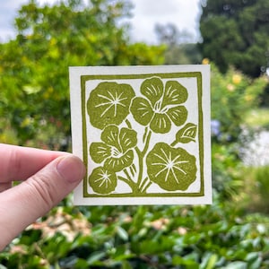May include: A square, off-white print featuring a green floral design. The design includes several stylized flowers and leaves, all in a solid, olive-green color. The print is held up against a blurred green background.