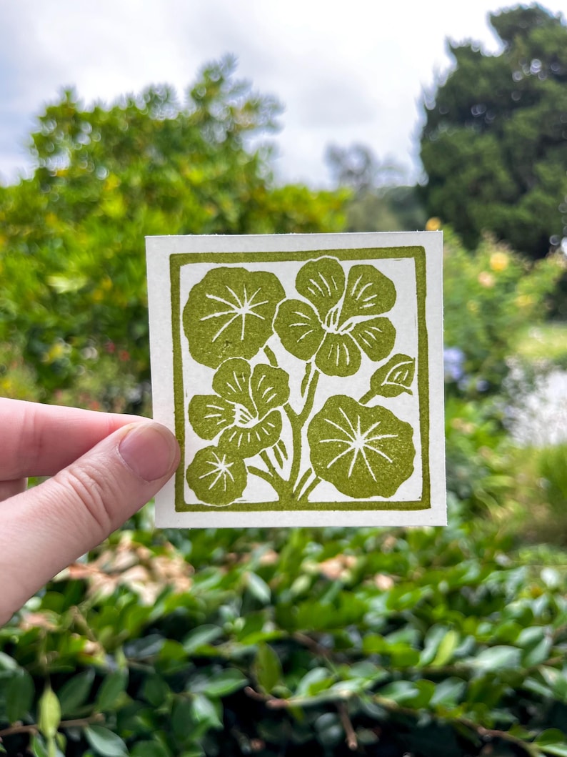 May include: A square, off-white print featuring a green floral design. The design includes several stylized flowers and leaves, all in a solid, olive-green color. The print is held up against a blurred green background.
