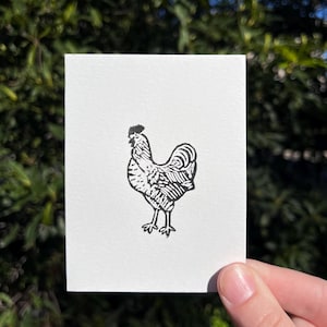 May include: A black and white rooster illustration on a white card. The rooster is depicted in a detailed linocut style, with textured feathers and a prominent comb. The card is held against a backdrop of green foliage.