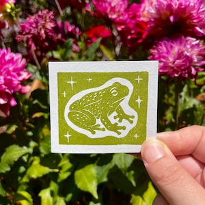 May include: A square print on white paper featuring a green block-printed illustration of a frog centered within a green border. The frog is surrounded by small white four-pointed star shapes. The design has a textured, hand-stamped appearance.