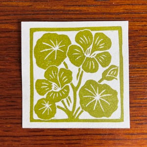 May include: A square, white card with a green floral design. The design features stylized flowers and leaves within a green border. The image is a linocut print, with a handmade aesthetic.