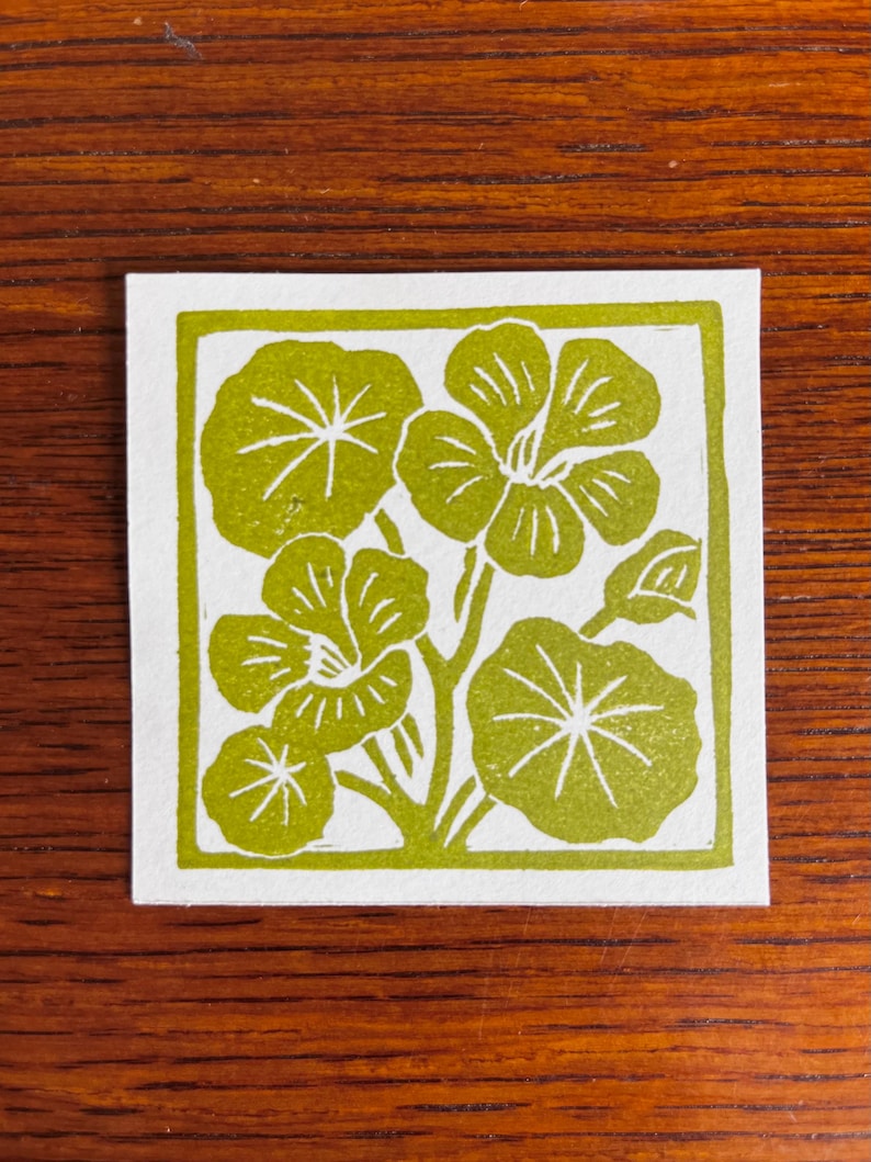 May include: A square, white card with a green floral design. The design features stylized flowers and leaves within a green border. The image is a linocut print, with a handmade aesthetic.