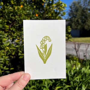 May include: A white card featuring a green illustration of a lily of the valley plant. The image shows the plant's bell-shaped flowers and long leaves. The card is held in front of a blurred background of greenery and a blue sky.
