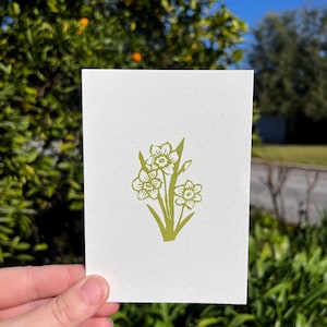 May include: A white card featuring a green floral design of daffodils. The illustration is a simple line drawing of the flowers and stems. The card is held in front of a blurred background of green foliage and a blue sky.